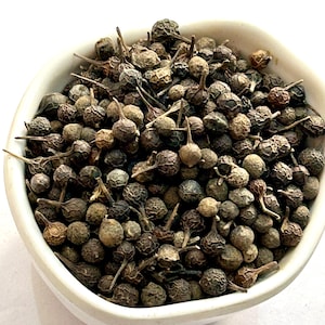 Op de afbeelding: Een witte kom gevuld met gedroogde, donkerbruine peperkorrels. De peperkorrels hebben een textuur en kleine steeltjes. De kom is wit en heeft een licht gebogen vorm. De peperkorrels zijn de belangrijkste focus van de afbeelding.
