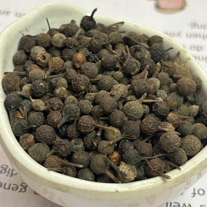 Op de afbeelding: Een witte keramische kom gevuld met donkerbruine, gedroogde peperkorrels. De peperkorrels hebben een textuur en kleine steeltjes. De kom staat op een bedrukte papieren achtergrond.