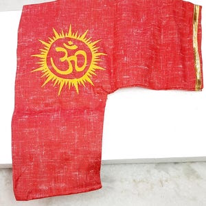 May include: Red garment featuring a yellow sun and Om symbol print. It has long sleeves and a gold trim. The fabric has a textured appearance. The garment is likely part of a traditional outfit.