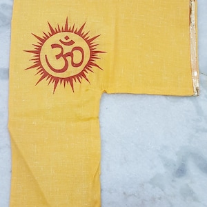 May include: A yellow garment featuring a red Om symbol within a sun design. The sleeve edge has a gold trim. The fabric is likely a lightweight linen or cotton. The garment is likely a religious item.