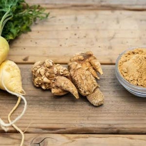 May include: A wooden surface showcases fresh maca root vegetables with green tops, whole maca roots, and a small glass bowl of maca powder. The maca roots are light yellow and tan in colour.