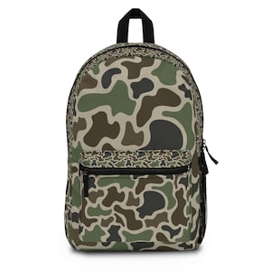 May include: A backpack featuring a camouflage pattern in shades of olive green, dark brown, and tan. The bag includes a large main compartment, a smaller front zippered pocket, a black top handle, and a black mesh side pocket for storage.