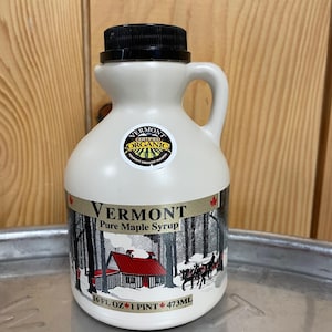 May include: A cream-colored Vermont Pure Maple Syrup bottle with a black cap and handle. The label features a winter scene with a red-roofed cabin and horse-drawn carriage. The bottle contains 16 fl oz (473ml) of syrup.