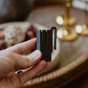 May include: A matte black plastic clip with a rectangular body and a vertical integrated hook. The object features a hollow centre section with three distinct vertical channels along the front face and a sturdy side-mounted tension clip.