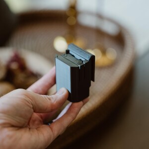 May include: A small, rectangular, matte black plastic component featuring a flat front surface and a top-mounted clip mechanism. The object has a structured, industrial design with visible seams and a solid, opaque finish, held in a vertical orientation.