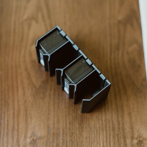 Peut inclure: Un organisateur noir rectangulaire &agrave; compartiments multiples, dot&eacute; d'une s&eacute;rie d'ailettes verticales et de fentes ouvertes. La structure est compos&eacute;e d'un mat&eacute;riau rigide au fini mat avec un motif g&eacute;om&eacute;trique r&eacute;p&eacute;titif pour les objets.