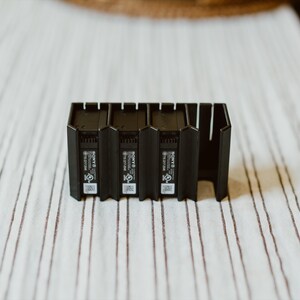 Peut inclure: Une station de charge en plastique noir dot&eacute;e de trois emplacements verticaux pour batteries d'appareil photo Sony. L'unit&eacute; pr&eacute;sente une base rectangulaire avec des ailettes de refroidissement et le logo Sony, des certifications et un QR code.