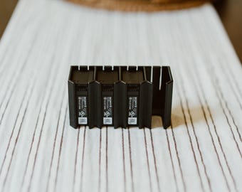 May include: A black plastic charging dock featuring three vertical slots for Sony camera batteries. The unit has a rectangular base with integrated cooling fins and clearly visible Sony branding, safety certification logos, and a QR code on each battery.