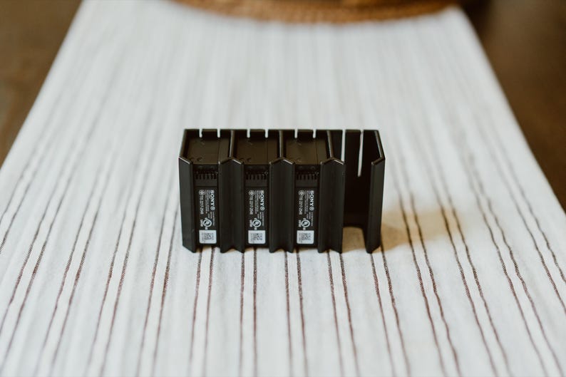 Peut inclure: Une station de charge en plastique noir dot&eacute;e de trois emplacements verticaux pour batteries d'appareil photo Sony. L'unit&eacute; pr&eacute;sente une base rectangulaire avec des ailettes de refroidissement et le logo Sony, des certifications et un QR code.