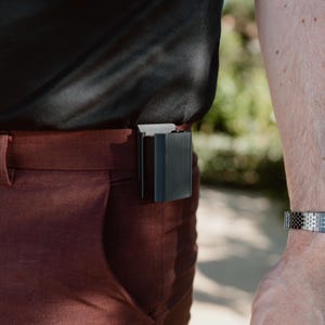 May include: A black rectangular clip-on accessory attached to the waistband of rust-coloured trousers. The device features a smooth, matte finish and a sturdy integrated clip mechanism that secures it firmly to the fabric of the trousers.