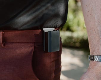 May include: A black rectangular clip-on accessory attached to the waistband of rust-colored trousers. The device features a smooth, matte finish and a sturdy integrated clip mechanism that secures it firmly to the fabric of the pants.