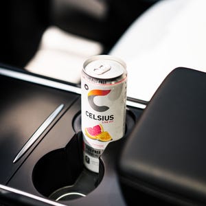 Peut inclure: Une canette de boisson &eacute;nergisante Celsius dans le porte-gobelet d'une voiture. La canette est blanche avec le logo Celsius et des graphiques de fruits. Le texte "Fizz-Free" et "Live Fit" est visible. Un stylo argent&eacute; est pos&eacute; &agrave; c&ocirc;t&eacute; du porte-gobelet.
