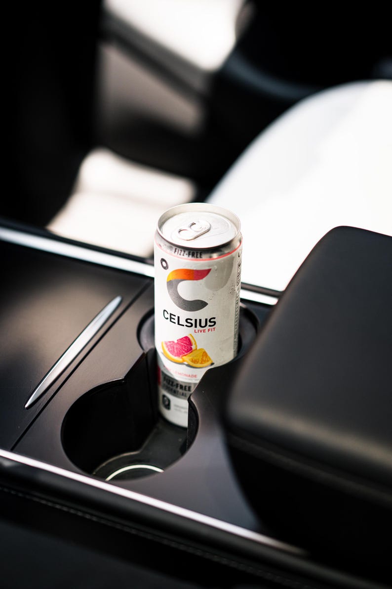 Peut inclure: Une canette de boisson &eacute;nergisante Celsius dans le porte-gobelet d'une voiture. La canette est blanche avec le logo Celsius et des graphiques de fruits. Le texte "Fizz-Free" et "Live Fit" est visible. Un stylo argent&eacute; est pos&eacute; &agrave; c&ocirc;t&eacute; du porte-gobelet.