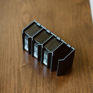 Peut inclure: Un support de rangement en plastique noir con&ccedil;u pour maintenir quatre batteries rectangulaires en position verticale. Le support pr&eacute;sente des fentes individuelles avec des c&ocirc;t&eacute;s ouverts et un cadre rigide angulaire, avec le marquage CE visible.