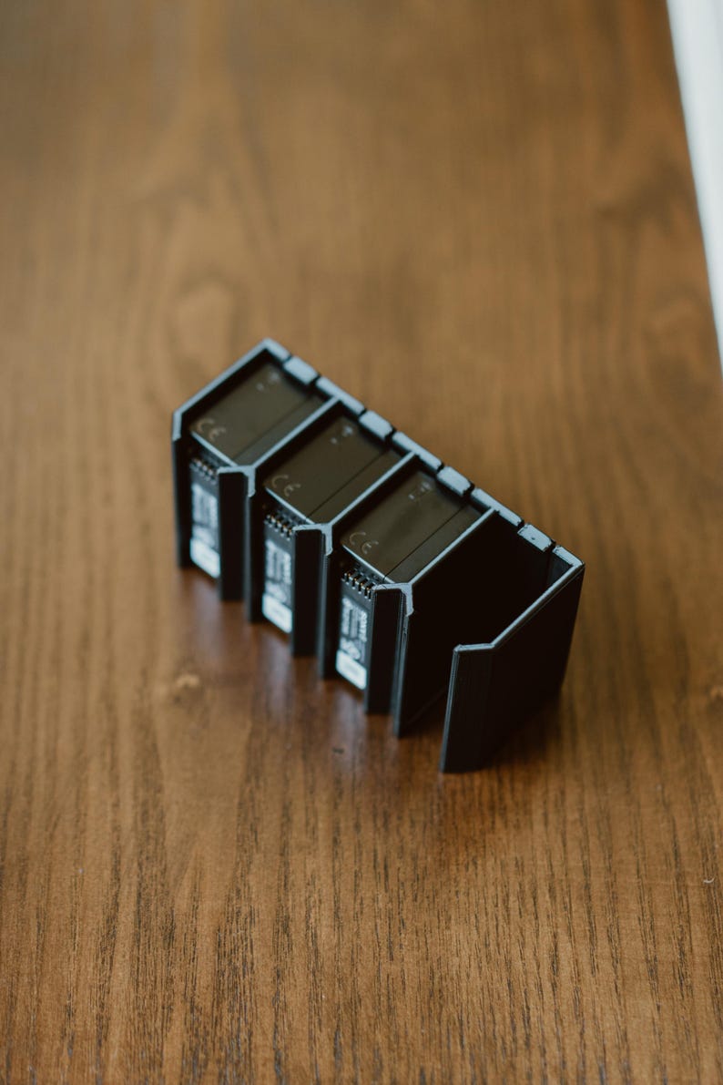 Peut inclure: Un support de rangement en plastique noir con&ccedil;u pour maintenir quatre batteries rectangulaires en position verticale. Le support pr&eacute;sente des fentes individuelles avec des c&ocirc;t&eacute;s ouverts et un cadre rigide angulaire, avec le marquage CE visible.