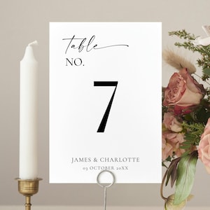 May include: A white table number card with the number 7 in black, displayed on a silver holder. The card also includes the text "Table No." and names. A white candle in a gold holder and a floral arrangement are in the background.