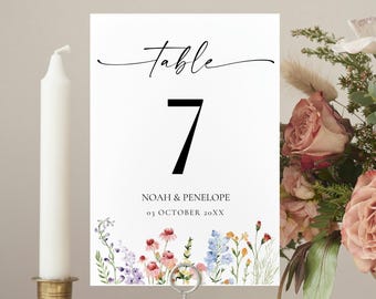 May include: A white table number card with the word "table" in script and the number 7 in black. The card has a floral border and is held in a metal stand. A white candle in a gold holder and a floral arrangement are also in the image.