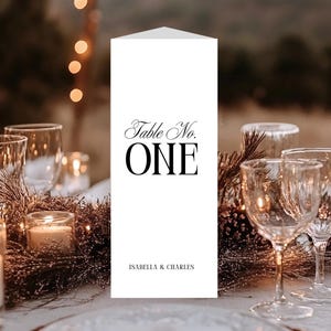 May include: A white table number card with the text "Table No. ONE" in black script and block letters. The card is on a table set with glassware, candles, and decorative greenery. The names "ISABELLA & CHARLES" are printed at the bottom.