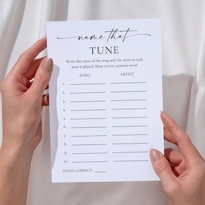 May include: A white paper game sheet with the text "name that TUNE" in elegant script. The sheet has spaces for song titles and artists, held by hands against a white fabric backdrop. The game is designed for a party or event.