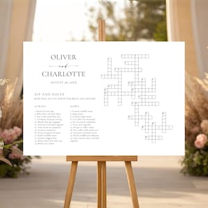 Peut inclure: Un panneau blanc sur un chevalet en bois affiche un jeu de mots croisés avec les noms "Oliver" et "Charlotte" et la date du 26 août 20XX. Le panneau indique "Sip and Solve" et comprend des indices. Des compositions florales et des éléments décoratifs entourent le panneau.