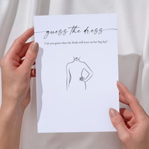 Pode incluir: Um cartão branco com o texto "guess the dress" e um desenho de uma figura feminina. O cartão é segurado por duas mãos com unhas cuidadas contra um fundo de tecido branco. O texto abaixo do título diz: "Can you guess what the Bride will wear on her big day?"