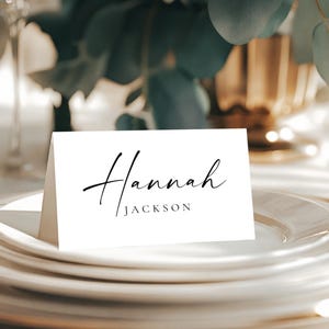 May include: A white place card with the name "Hannah Jackson" in elegant black script sits atop a stack of white plates. The card is angled slightly, with a blurred background of greenery and gold accents.