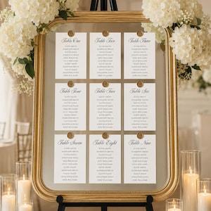 Puede incluir: Un plano de mesa sobre espejo con marco dorado que muestra nueve tarjetas blancas etiquetadas de Table One a Table Nine. Cada tarjeta presenta una lista de nombres en texto negro y está fijada al cristal con un sello de cera dorado.