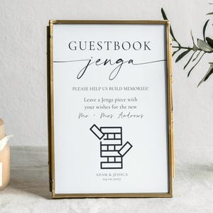 Puede incluir: Un letrero enmarcado con las palabras "GUESTBOOK jenga" en escritura elegante. El letrero anima a los invitados a dejar deseos en piezas de Jenga. Una vela y vegetación están en el fondo.