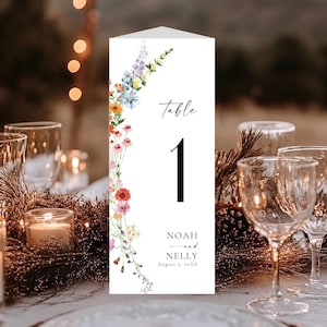 May include: A white table number card with floral accents and the number 1. The card features the word "table" in script, along with the names "Noah and Nelly" and a date. The card is surrounded by candles and glassware.