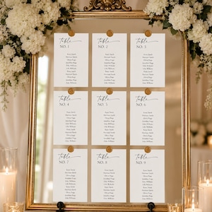 May include: A gold-framed seating plan with nine cards, each labelled "Table No." and listing names. The cards are held by gold pins. White floral arrangements and candles in glass holders decorate the background.