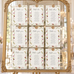 May include: A large, ornate gold-framed mirror displaying nine white rectangular seating chart cards arranged in a grid. Each card features floral borders, a table number, a list of names, and a gold wax seal at the top centre of each card.
