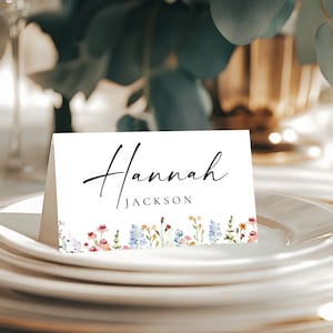 May include: A white place card with the name "Hannah Jackson" in elegant black script. The bottom edge features a watercolour floral design in shades of pink, blue, and yellow. The card rests on a stack of white plates, suggesting a wedding or event setting.