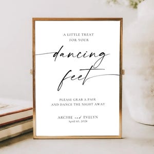 May include: A framed sign with gold trim and white background. The sign reads "A LITTLE TREAT FOR YOUR dancing feet" in elegant script. Below, it says "PLEASE GRAB A PAIR AND DANCE THE NIGHT AWAY." The bottom of the sign says "ARCHIE and EVELYN April 10, 2028."