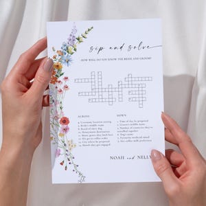 Pode incluir: Um cartão de jogo de casamento retangular branco intitulado Sip and Solve com um jogo de palavras cruzadas, apresentando uma borda floral em aquarela colorida no lado esquerdo. O texto inclui How well do you know the bride and groom e os nomes Noah and Nelly.
