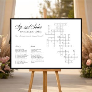 Puede incluir: Un crucigrama enmarcado sobre un caballete de madera, con el texto "Sip and Solve" y los nombres "Isabella & Charles". El rompecabezas está diseñado para una boda, con pistas relacionadas con los novios.