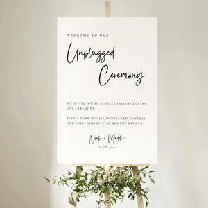 Può includere: Un cartello bianco su un cavalletto con le parole "Welcome to our Unplugged Ceremony" in elegante scrittura nera. Il cartello include anche una richiesta di spegnere telefoni e fotocamere. La parte inferiore del cartello è decorata con vegetazione.