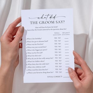 May include: A white paper game sheet titled "What Did The Groom Say?" with questions about the bride. The sheet is held by two hands against a white fabric background. The game is designed for a bridal shower.