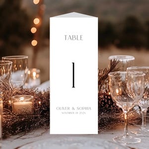 May include: A white table number card with the word "TABLE" at the top and the number "1" in the center. The names "OLIVER & SOPHIA" and the date "NOVEMBER 19 2025" are printed at the bottom. The card is on a table with glasses and candles.