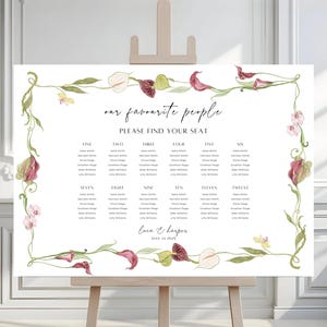 Puede incluir: Un cartel rectangular blanco de distribución de asientos para bodas sobre un caballete de madera, con un borde floral de acuarela de calas rojas y blancas. El texto dice: our favourite people, PLEASE FIND YOUR SEAT, mesas del uno al doce, y Lace & Harper MAY 14 2024.