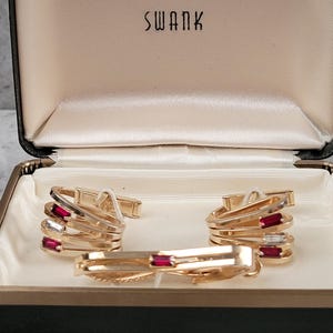 May include: A gold-tone jewelry set consisting of two cufflinks and a tie bar, presented in a black Swank box with a cream interior. Each piece features a multi-layered design accented with rectangular red and clear stones set in gold-tone metal.