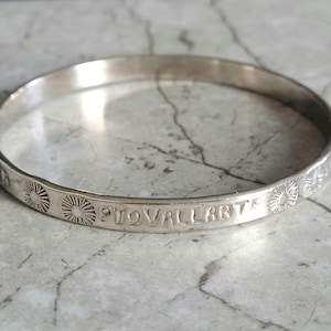 May include: A silver-toned metal bangle bracelet featuring a smooth, polished finish. The exterior surface is engraved with the text PTO VALLARTA and is decorated with repeating sunburst patterns spaced evenly around the circumference of the band.