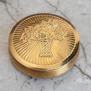 Puede incluir: Un estuche compacto redondo y dorado con un diseño detallado de cesta floral. La tapa presenta un relieve en relieve de rosas y otras flores dentro de una cesta tejida, rodeada de líneas radiales. El compacto está sobre un fondo de mármol.