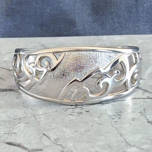 May include: A silver-toned cuff bracelet with an intricate openwork design. The bracelet features a textured central panel and decorative swirling patterns along the edges. The metal has a polished finish, reflecting light.