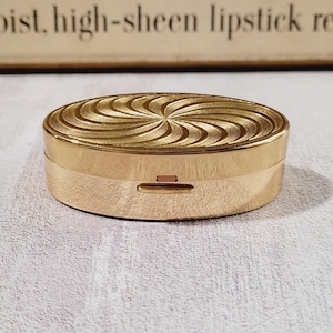 May include: A gold-colored, oval compact with a spiral design on the lid. The compact has a small rectangular clasp on the front. The background is a light, textured surface.