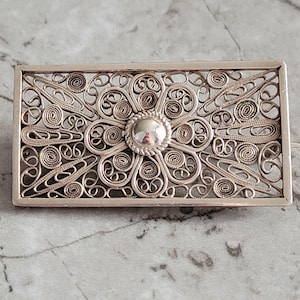 May include: A rectangular silver brooch with an intricate filigree design. The brooch features a central flower-like element surrounded by swirling patterns and a raised, circular center. The metalwork is detailed and delicate, showcasing a vintage aesthetic.