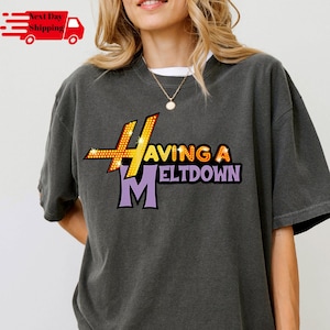 Pode incluir: T-shirt cinza escuro com o texto "Having a Meltdown" em uma fonte colorida e estilizada. O design apresenta um grande "H" e "M" com a palavra "Having" em amarelo e "Meltdown" em roxo. A camisa é de estilo casual.