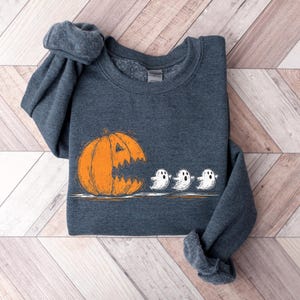 Può includere: Una felpa grigio scuro con una grafica a tema Halloween. Il design include una grande zucca arancione con una bocca frastagliata e tre piccoli fantasmi bianchi. Il design è giocoso e festoso.