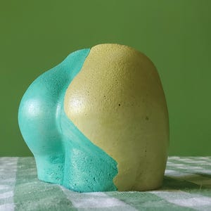 May include: An abstract sculpture with a rounded shape, painted in teal and yellow. The sculpture is set against a green backdrop and rests on a green and white checkered surface. A modern art piece.