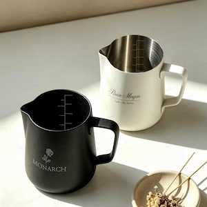 Könnte beinhalten: Ein schwarzer Milchkännchen aus Edelstahl mit einer Rosengrafik und dem Text MONARCH sowie ein weißes Milchkännchen aus Edelstahl mit dem Text Briar Morgan Light Like Aurora. Beide Kännchen verfügen über interne Messmarkierungen.