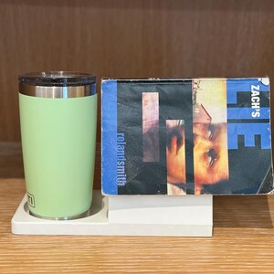 May include: A light green Yeti tumbler with a silver rim and a clear lid, next to a book titled "Zach's Lie" by roland smith. The book cover features a blue and black design with a person's face.